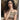 A woman with long, curly dark hair wearing a sheer, off-the-shoulder top with a brown and beige abstract pattern, posing outdoors next to a wooden post.